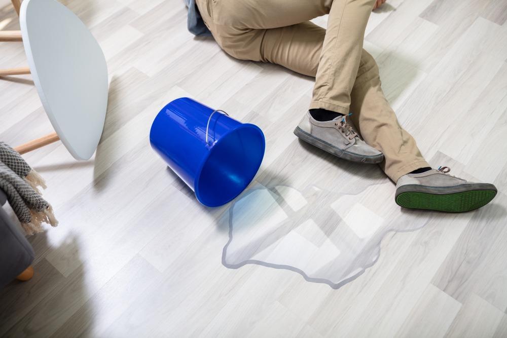 A man on the floor after slipping due to liquid from a spilled over bucket.