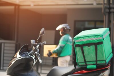 Man in green jacket walks away from a motorcycle with a food delivery parcel strapped to the back after a food delivery accident.