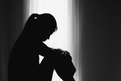 A woman huddles in the shadows of a dimly lit room. There are many common signs of sexual abuse, including physical, behavioral, social, and emotional.